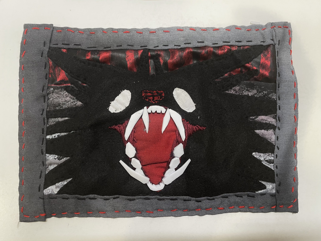 Textile artwork made up of red black grey and white fabric. Grey border with red and black stitches, and black center with wolf like face in middle, with red mouth and white teeth and eyes.