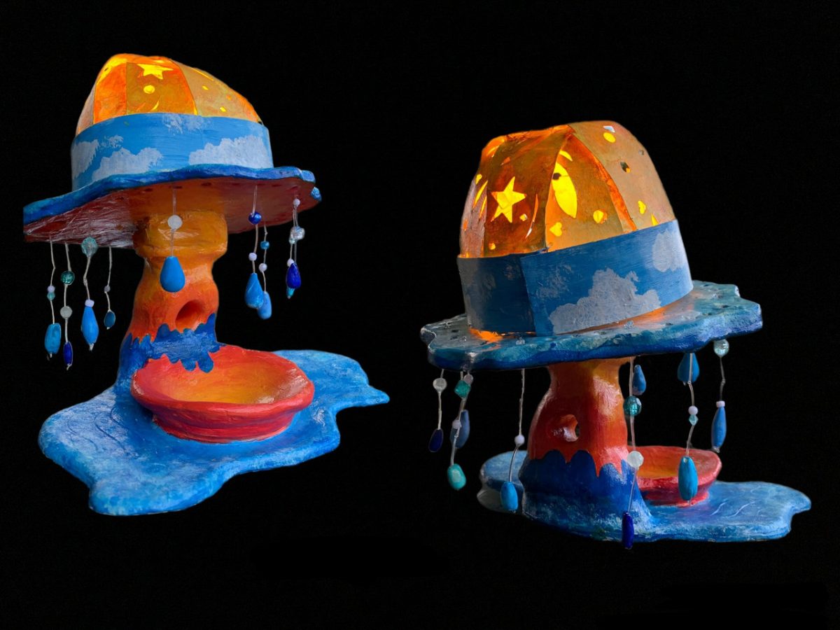 Photograph of two multi-colored sculptures shaped like mushrooms with rain coming down and puddles underneath. The sculptures are orange with stars on the top and underside, but blue with clouds on the rim and bottom.