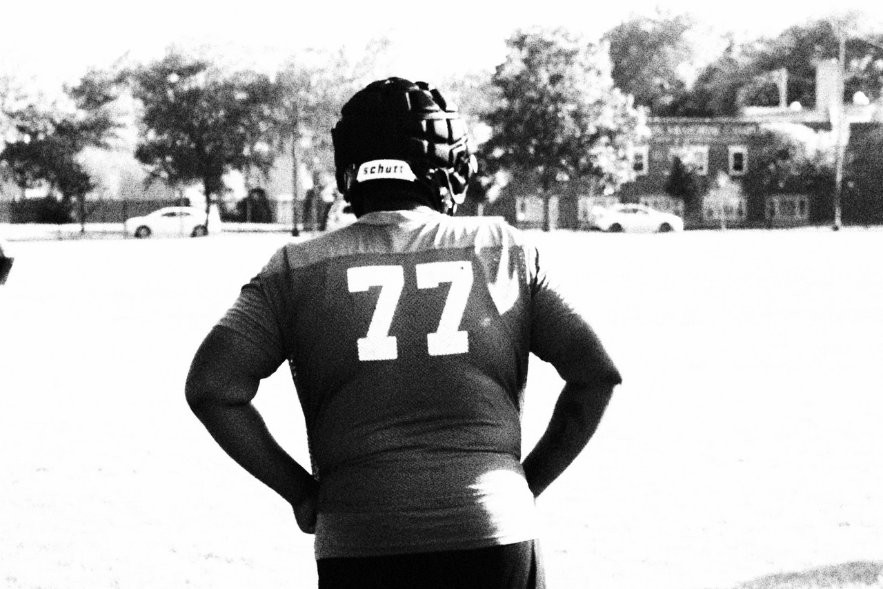black and white photograph of back of a person in jersey number 77 and helmet standing with hands on hips