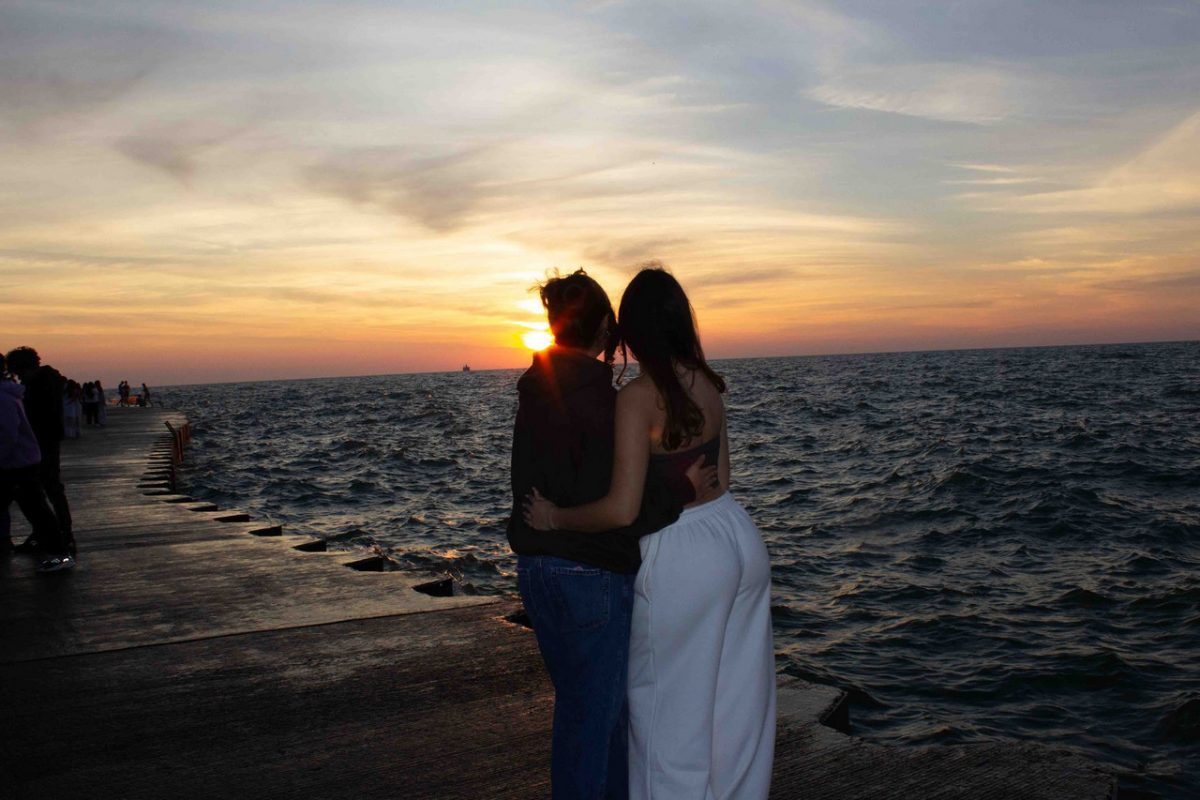 Photograph of two people standing with backs to camera in front of lake and sunrise, hugging.