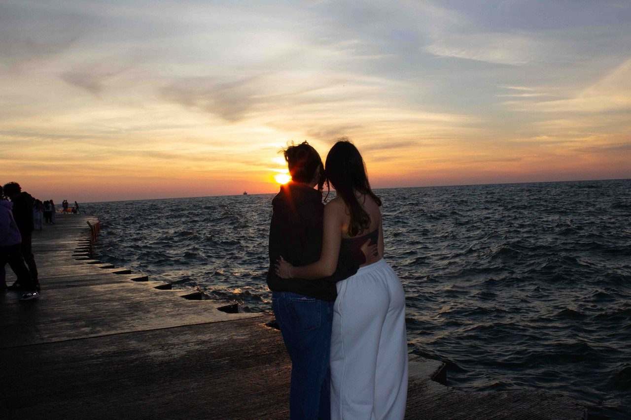 Photograph of two people standing with backs to camera in front of lake and sunrise, hugging.