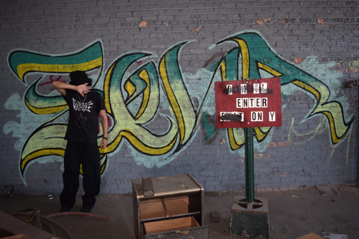 photograph of person standing in black shirt and pants in front of wall with graffiti and red sign