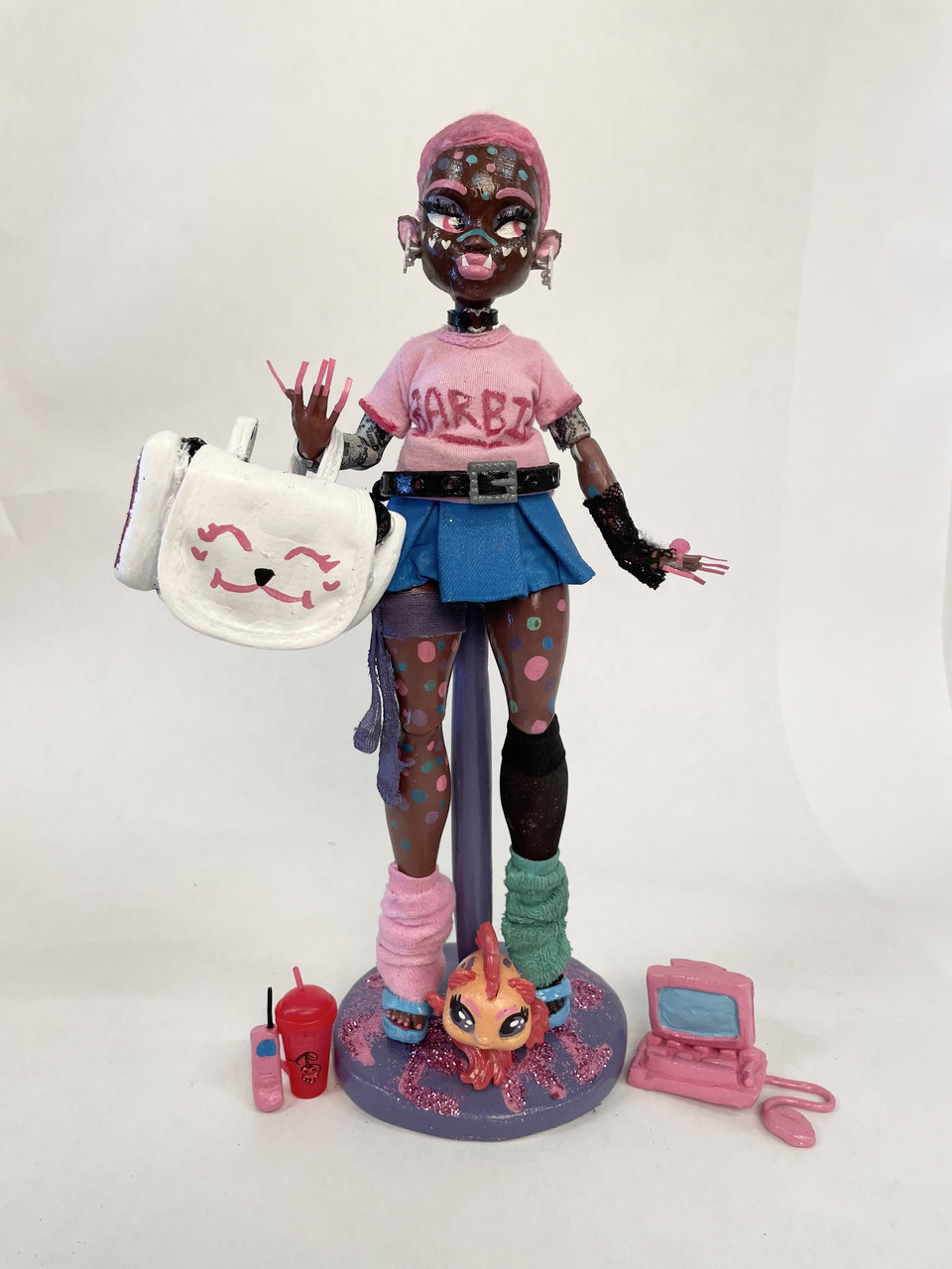 Photograph of doll sculpture with dark skin and large white bag. Doll is wearing pink shirt and denim skirt with accessories by their feet.