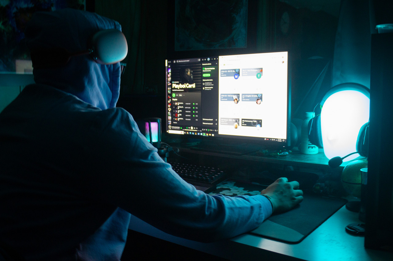 dark photograph of person with back to camera looking at computer screen wit headphones on and hand on mouse