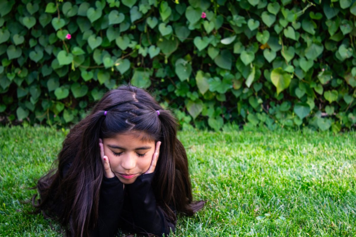 Photograph of dark haired younger girl laying in grass on stomach with head in hands and leaf covered background