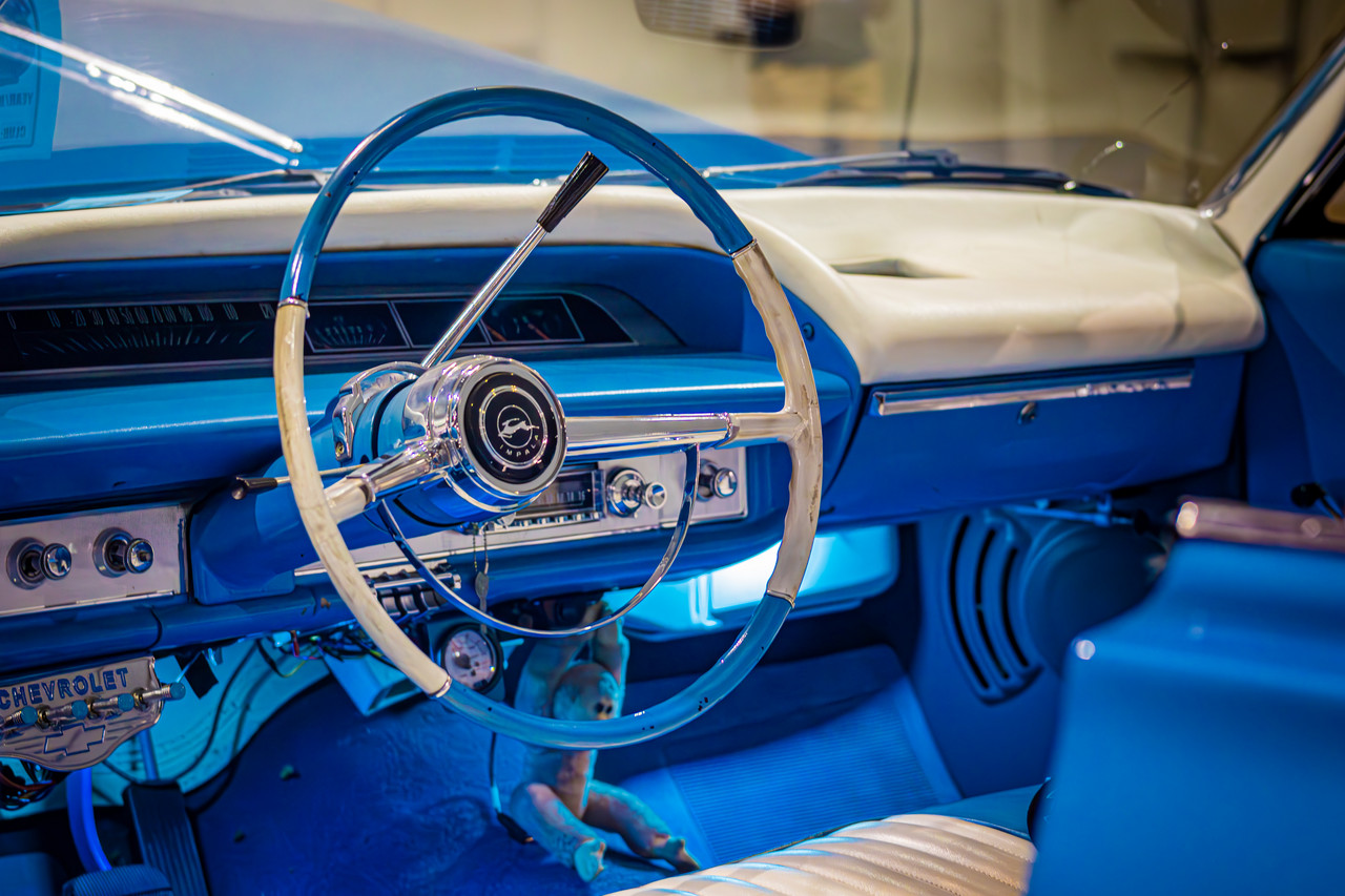 Photograph of inside of vintage car with blue and white interior, large steering wheel, and light under dashboard
